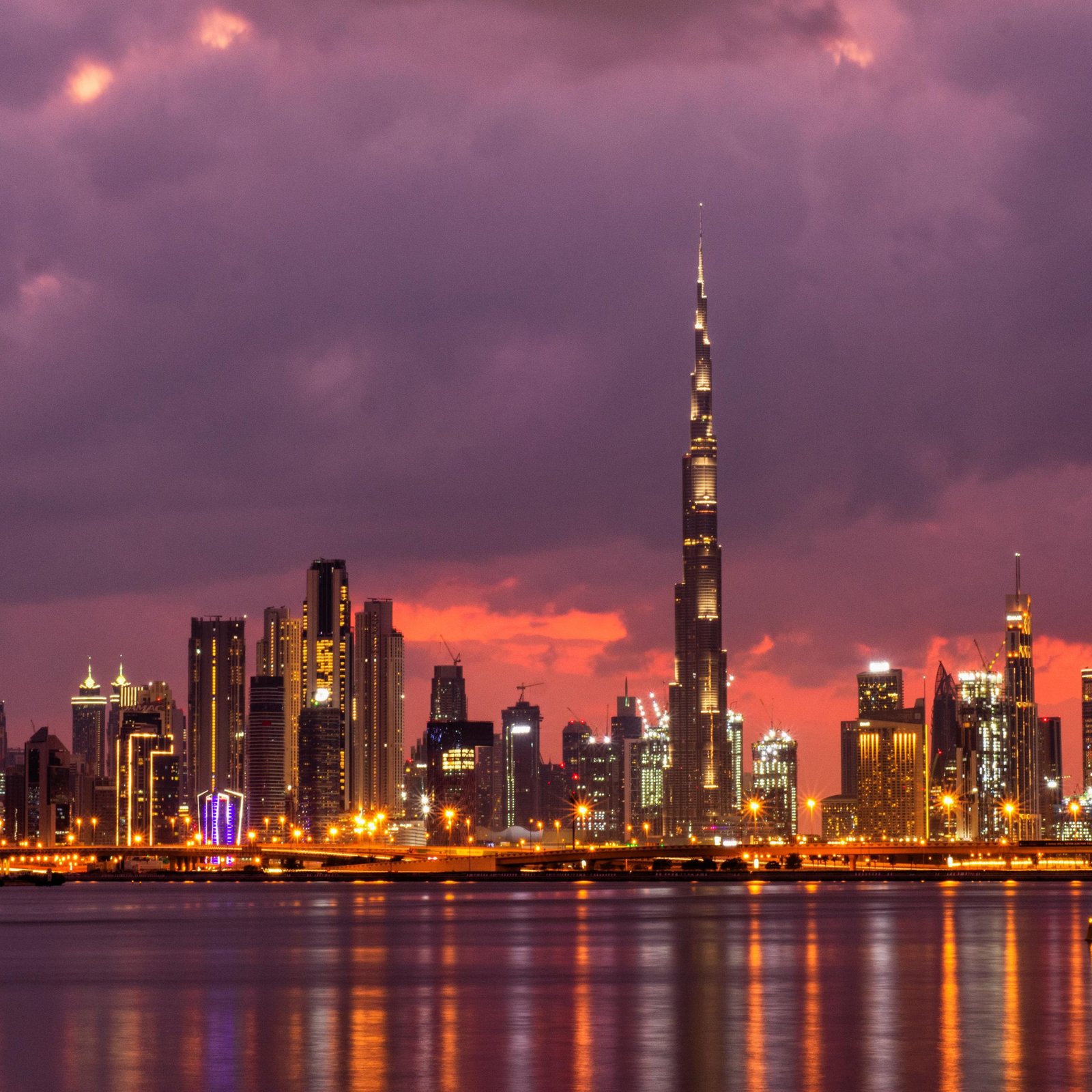 A stunning panoramic view of the Dubai skyline at sunset, featuring the illuminated Burj Khalifa and other skyscrapers reflecting on the water.